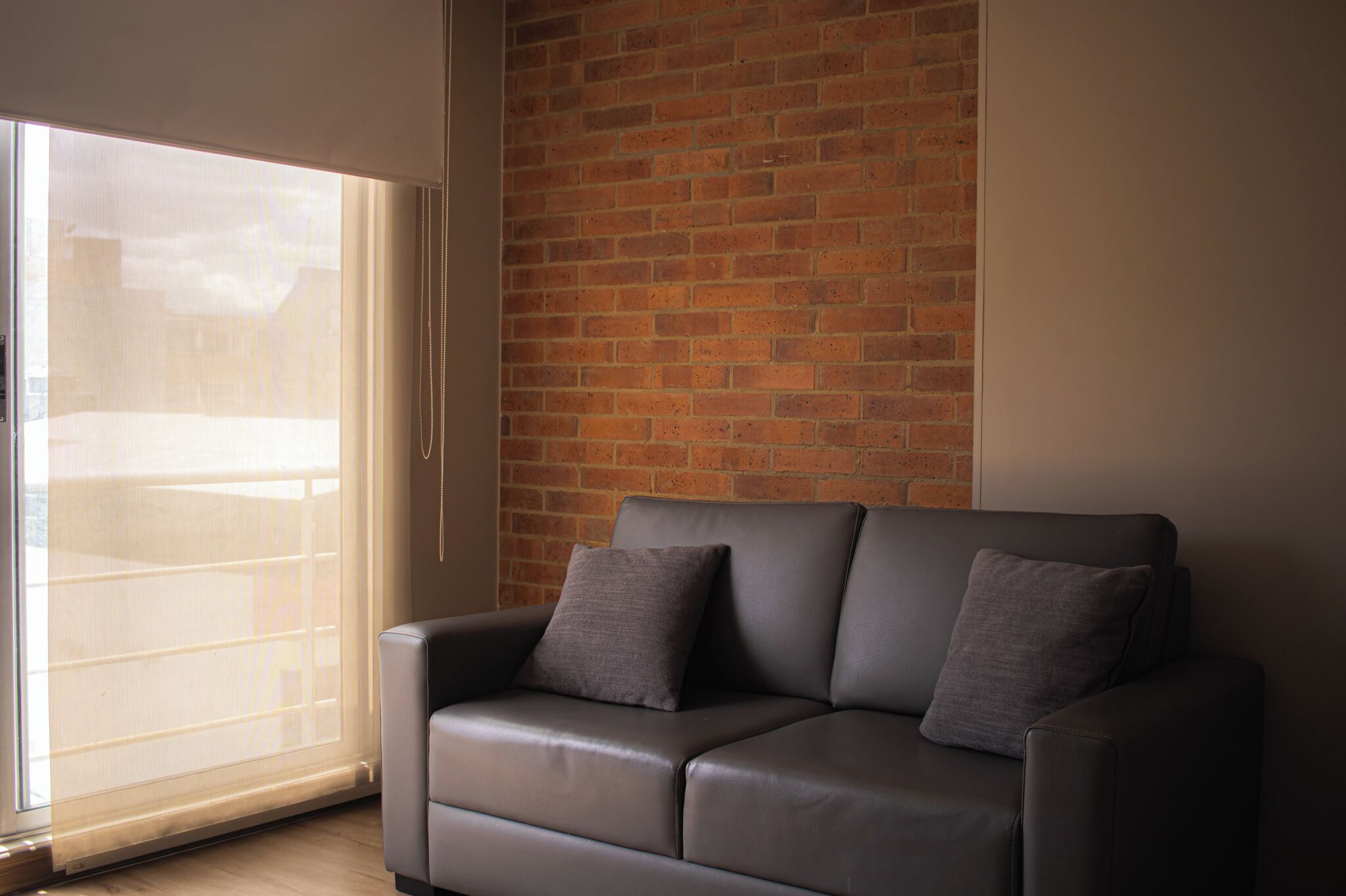 A modern living room with a leather sofa, two cushions, a brick wall, and a large sliding glass door.