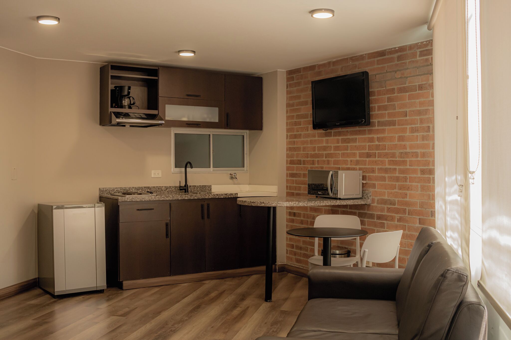 Cocina compacta con refrigerador, cafetera y microondas. Hay una zona de comedor con mesa y sillas, además de un sofá.