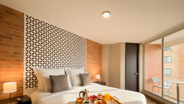 A hotel room with a large bed, a tray of breakfast, a brick wall, and a view of the outside. - Hemma Bogotá Chapinero Premium Suites Hotel (Bogotá)