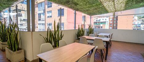 An outdoor patio with tables and chairs, surrounded by potted plants and a view of buildings.