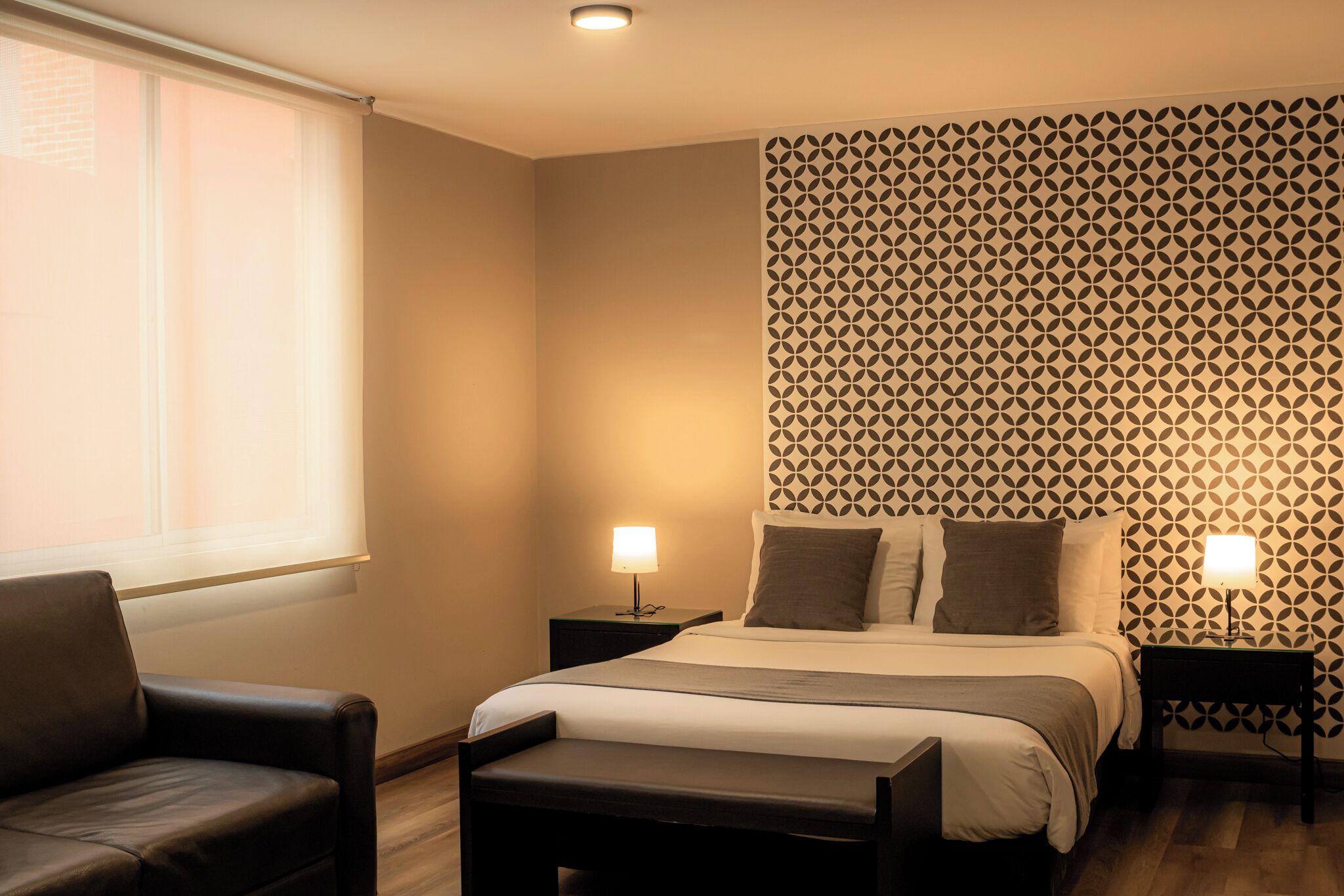 Quarto de hotel com cama, duas mesas de cabeceira com luminárias, uma cadeira e um banco.