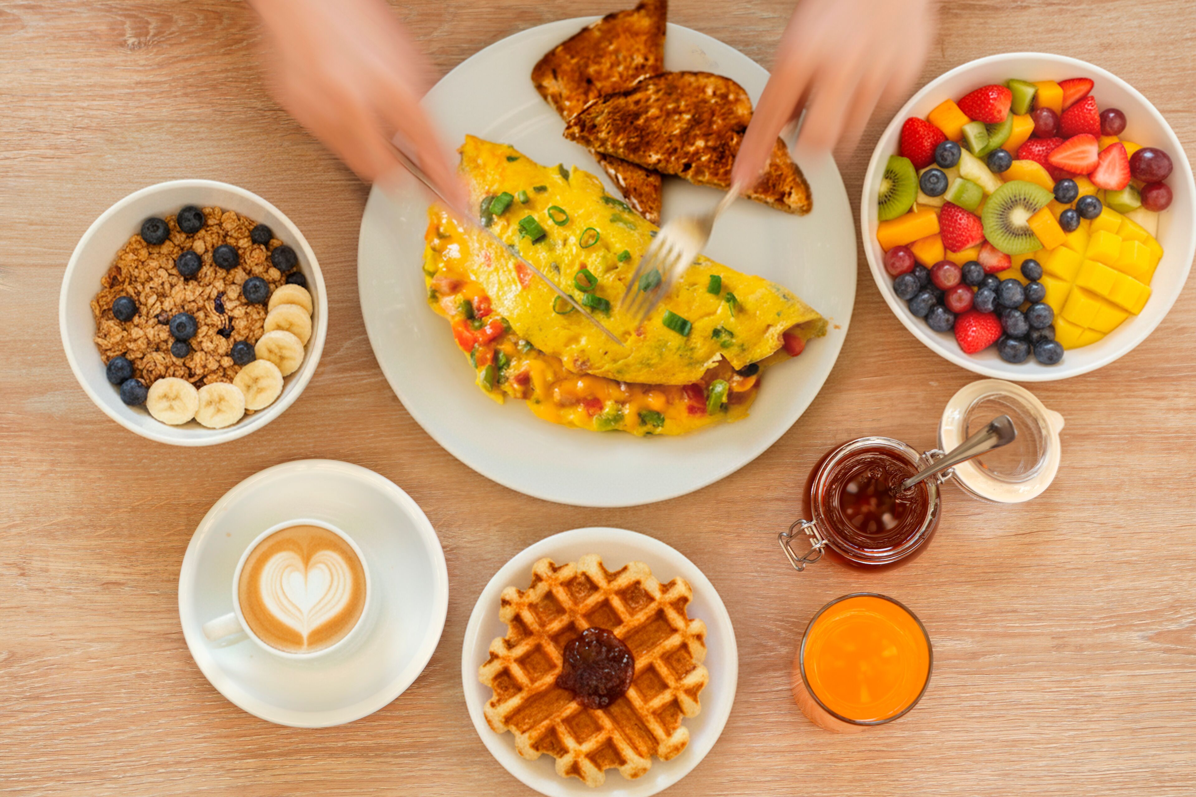 A breakfast setting with toast, scrambled eggs, fruit salad, granola with bananas, a waffle with jam, a coffee cup with latte art, and a glass of orange juice.