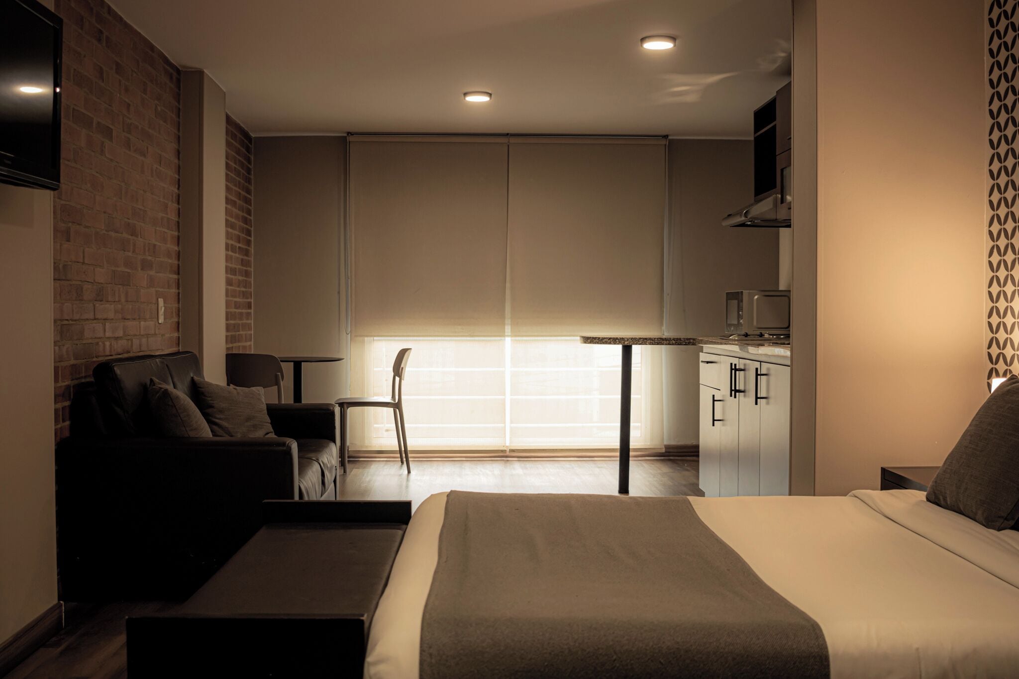 A modern hotel room with a bed, a seating area with a sofa and chairs, a small kitchenette, and a large window with blinds.