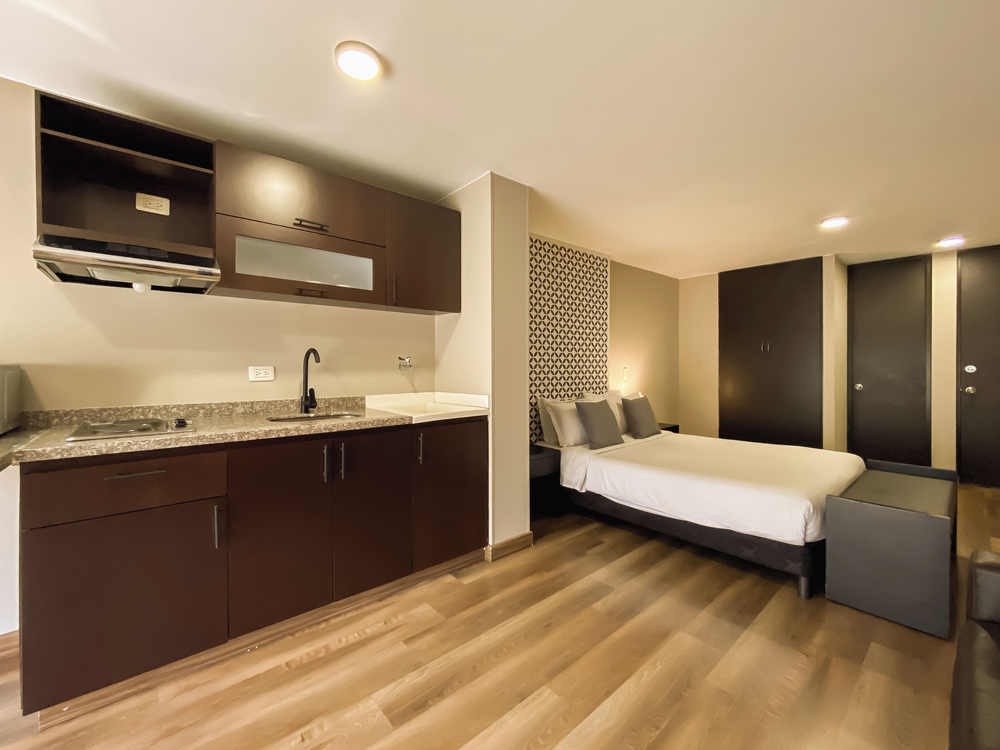 A compact hotel room with a kitchenette, a bed with a headboard, and a wardrobe.