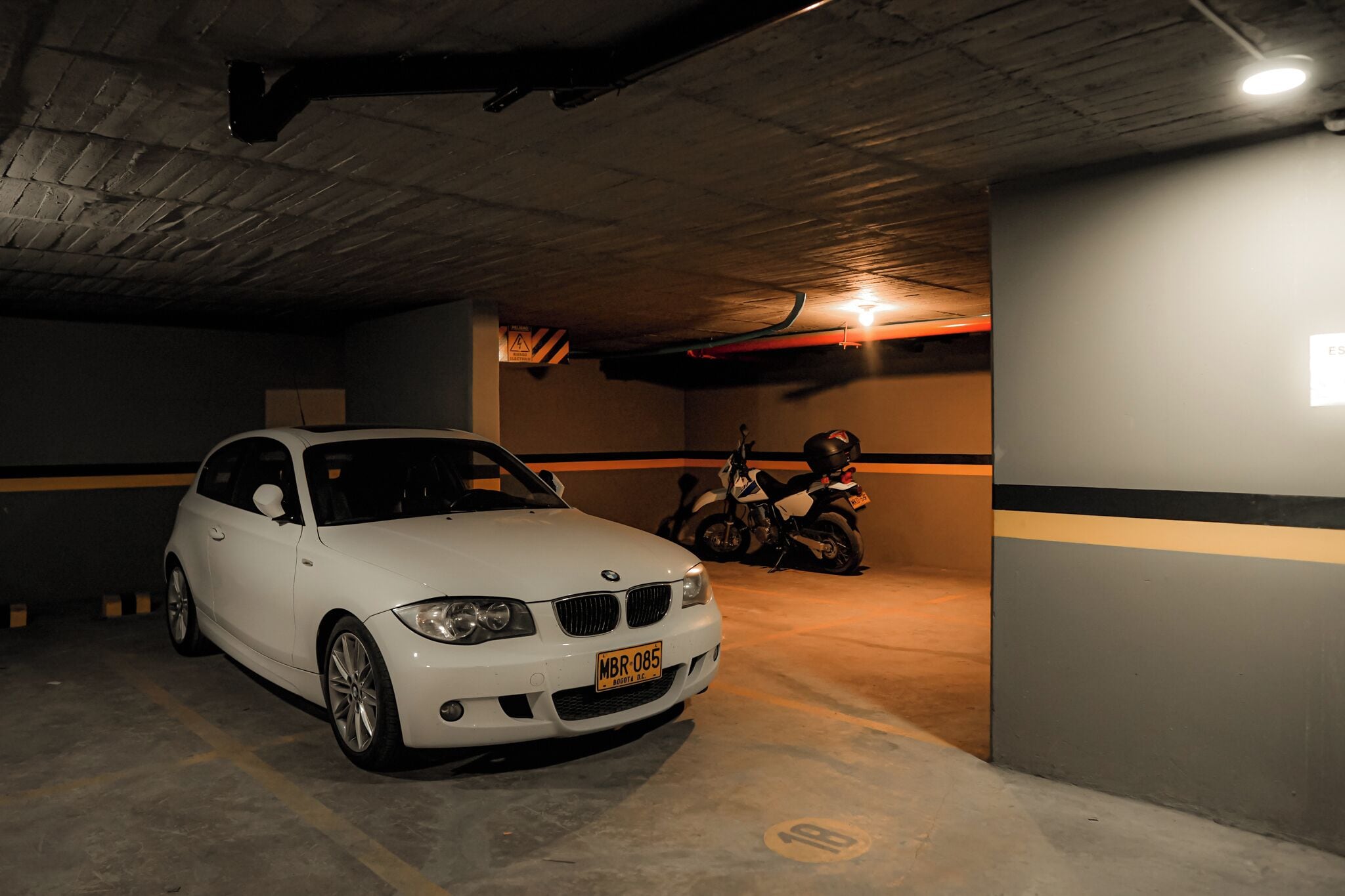 A white car parked in a dimly lit parking garage with concrete walls and a motorcycle in the background.