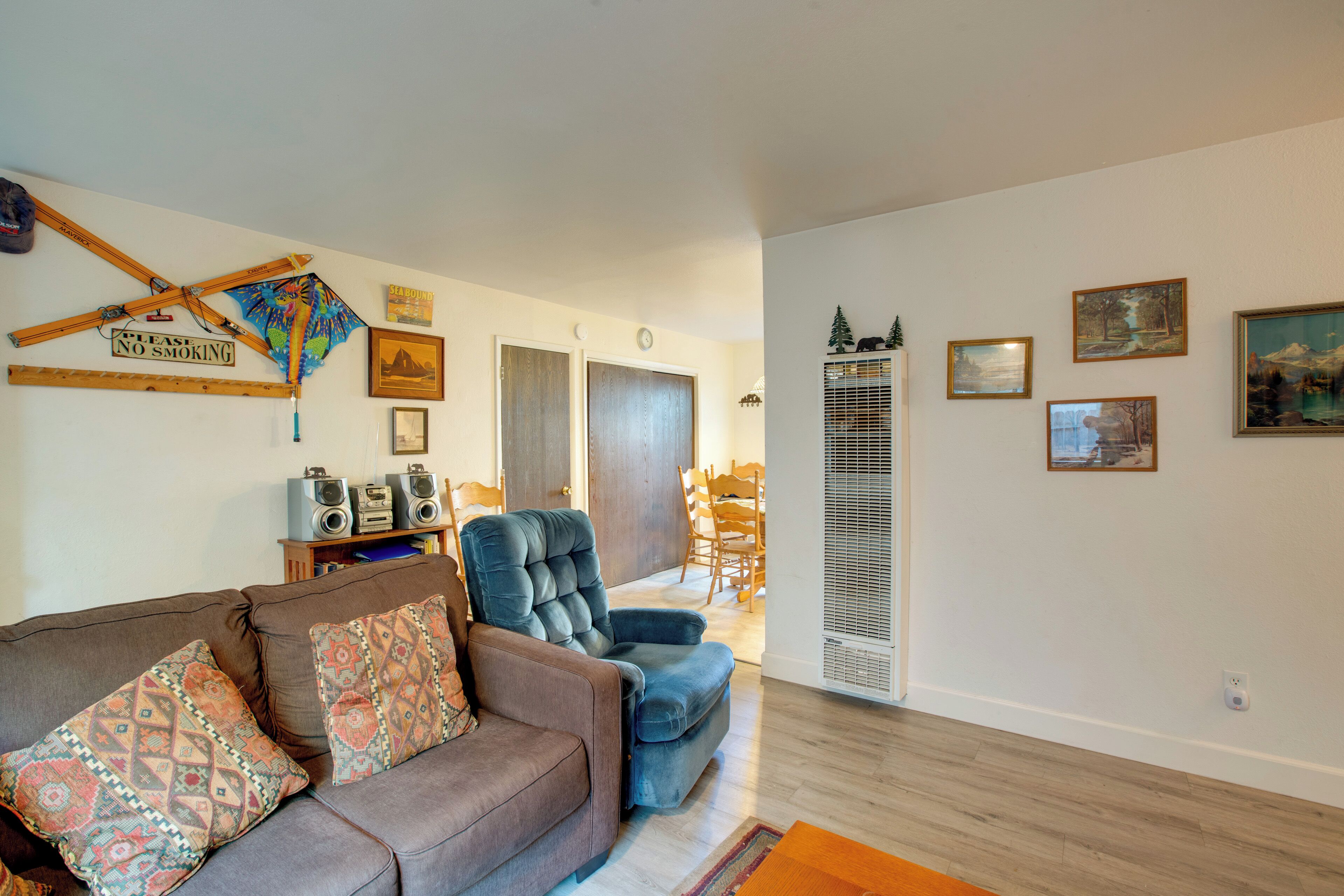 A living room with a brown sofa, a blue recliner, and a wooden coffee table. There are framed pictures on the wall and a 'No Smoking' sign.
