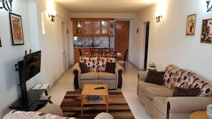 Una sala de estar con un sofá, una mesa de centro y un televisor. Se observa una cocina al fondo con armarios de madera y un comedor.