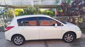 Un coche utilitario blanco estacionado sobre una superficie de hormigón, con una valla de malla metálica y vegetación al fondo.