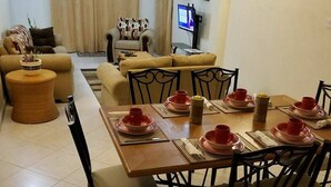 Un comedor con una mesa larga dispuesta para cuatro personas, rodeada de sillas. Se ve un sofá y una pequeña mesa redonda con una planta al fondo.