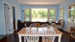 A dining area with a wooden table and white chairs, a sofa, and a view of a lake through large windows. - Lakeside cottage overhanging the water, beautiful window views across the lake (Ledyard)