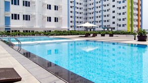 A swimming pool with lounge chairs and umbrellas, surrounded by a multi-story building with balconies. - M Place at Abs by Noemi (Quezon City)