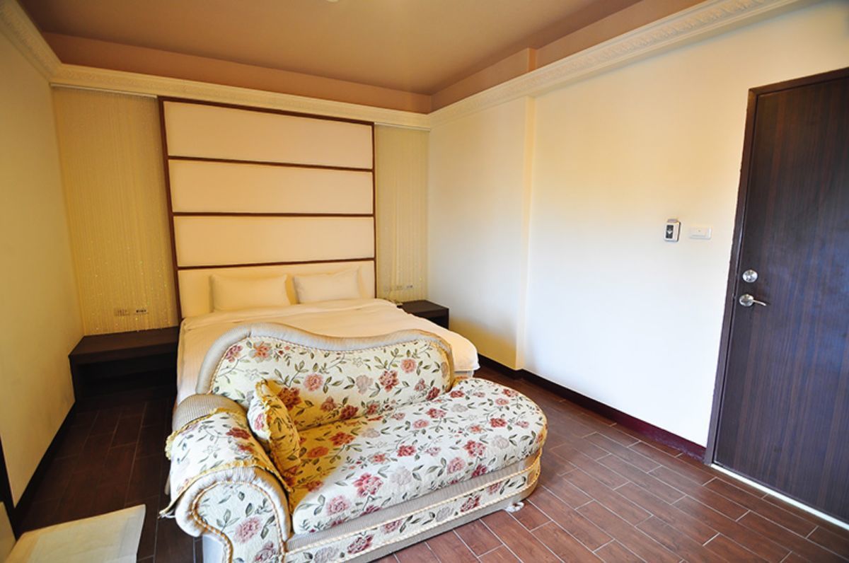 A bedroom with a bed, a floral sofa, a wooden floor, a door, and a wall with a light switch.