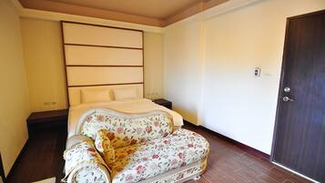 A bedroom with a bed, a floral sofa, a wooden floor, a door, and a wall with a light switch.