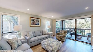 Un salon spacieux avec un canapé, un fauteuil et un pouf, un coin repas avec des chaises, et un balcon offrant une vue sur les arbres.