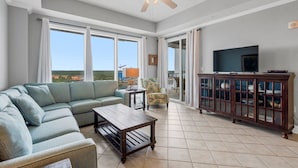 A spacious living room with a sectional sofa, a coffee table, a television on a cabinet, and large windows offering a view. - Waterfront Condo | Marina Views | Pools & Lazy River | On-Site Dining | Sleeps 8 (Orange Beach)
