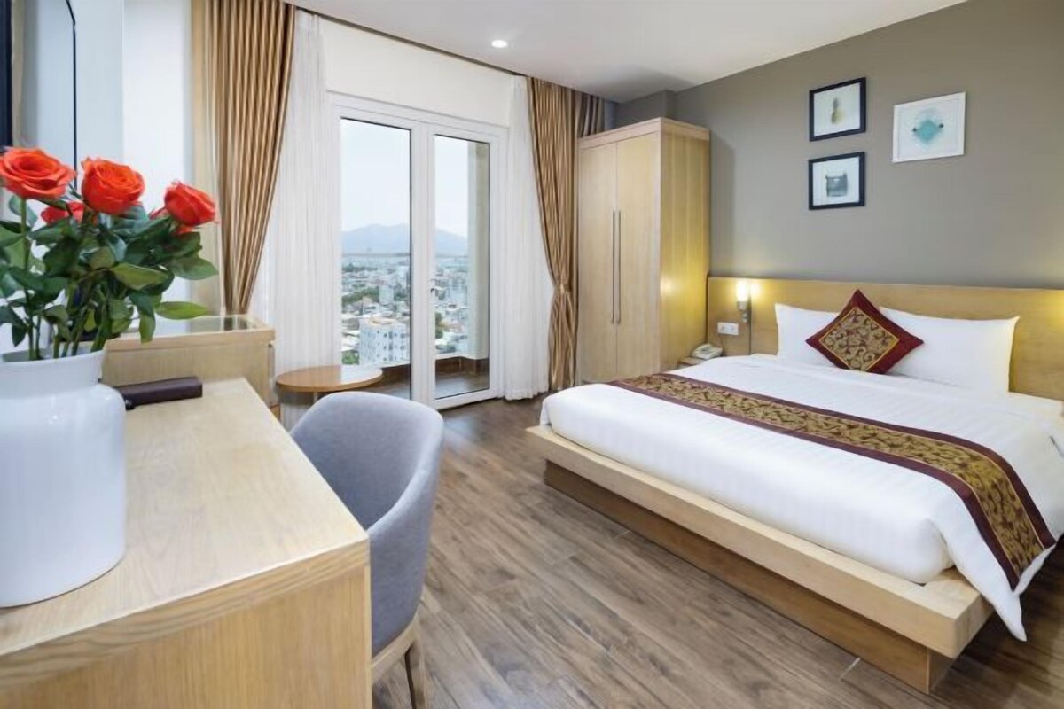 Habitació d'hotel amb un llit, una taula de treball, una cadira, un vas amb flors, vista a la ciutat i un balcó.