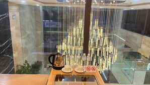 A modern hotel lobby with a glass coffee station, a decorative hanging light fixture, and a view of the cityscape through the glass.