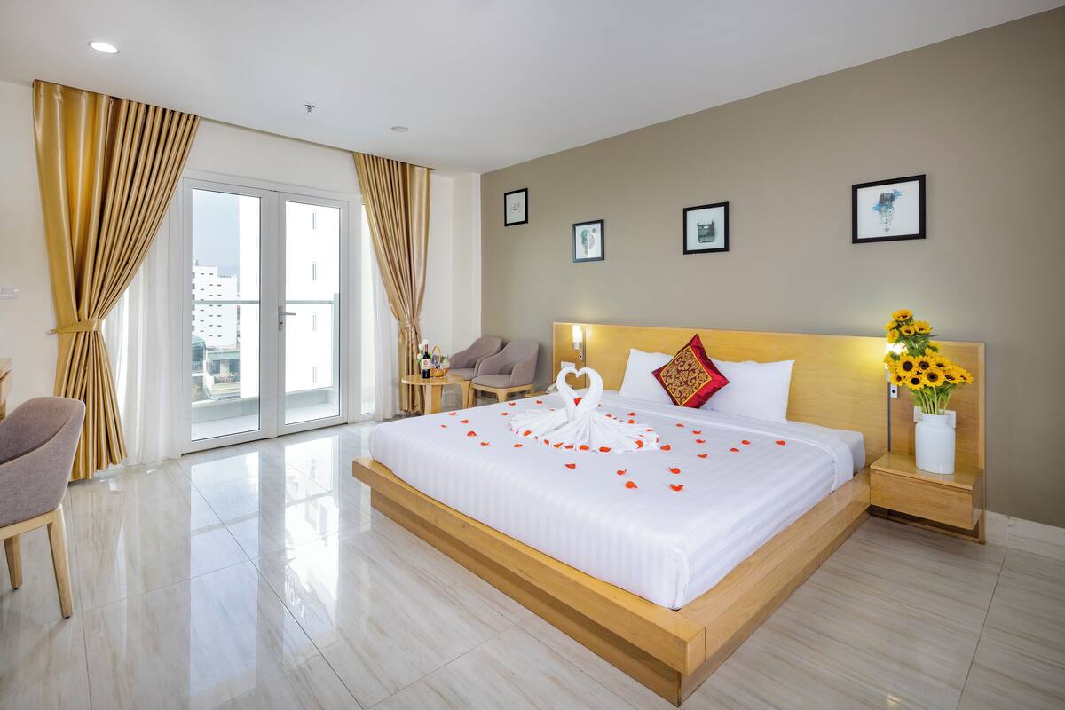 Une chambre d’hôtel avec un grand lit, une tête de lit en bois, une table de chevet ornée d’un vase de tournesols et une vue sur la ville depuis les portes du balcon.