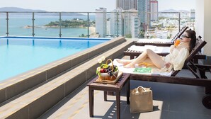 A poolside area with a view of the city, a lounge chair, a table with a basket of fruit, and a bag.