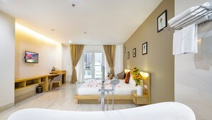 A modern hotel room with a large freestanding bathtub filled with rose petals, a bed with a view of the city, a desk with a chair, and a vanity with a mirror.