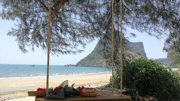 A beach scene with a swing set, a rocky outcrop, and a distant view of boats and a mountain.