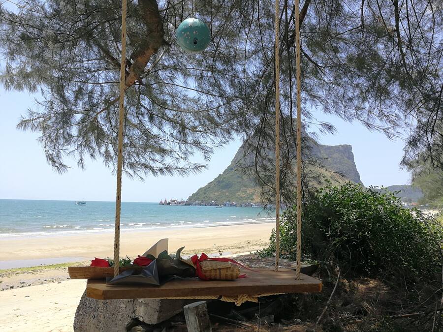 A beach scene with a swing set, a rocky outcrop, and a distant view of boats and a mountain.