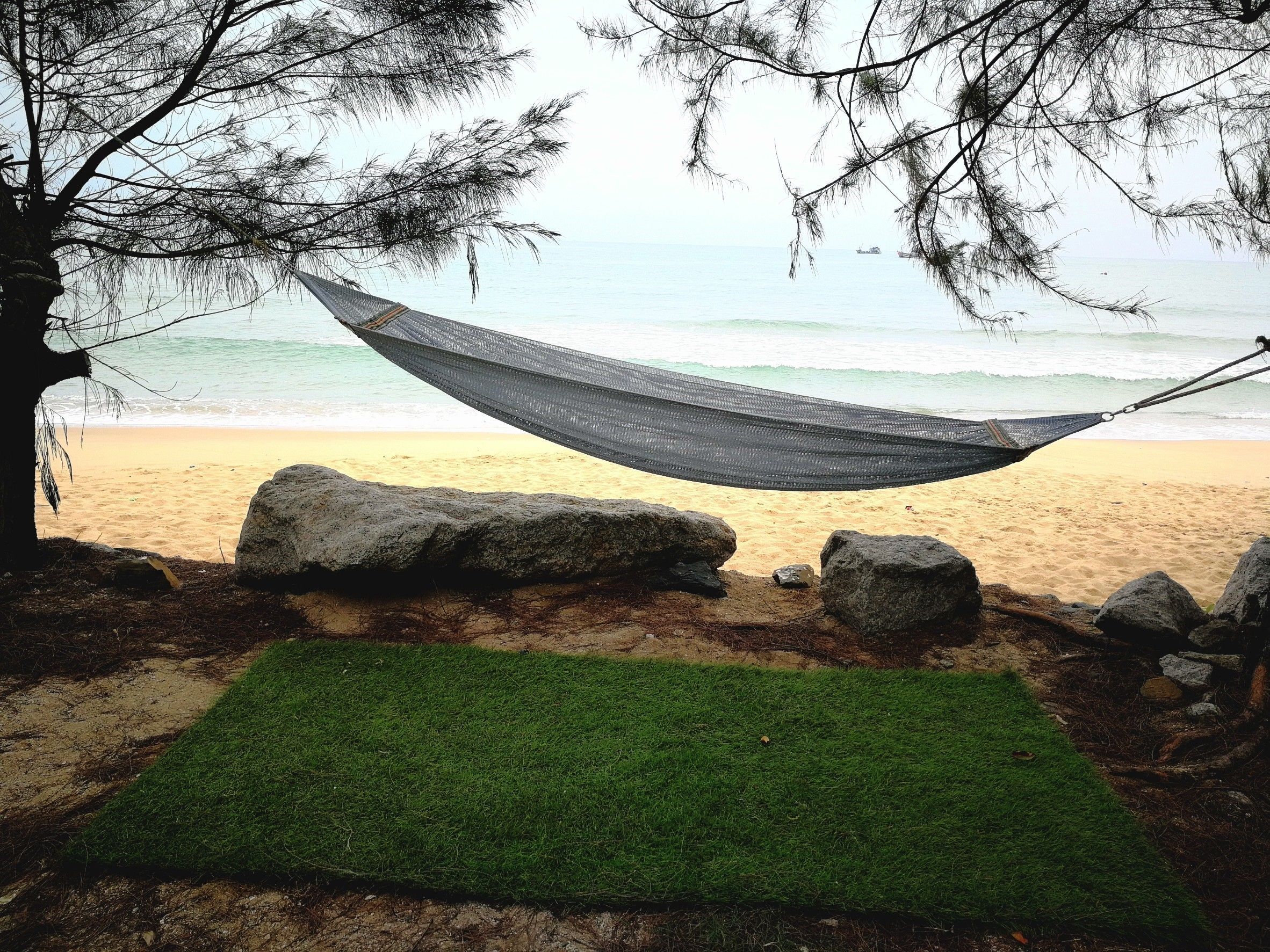 A hammock tied between two trees on a sandy beach with a green mat in front.