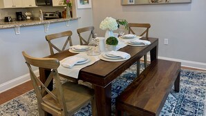 Un comedor con una mesa de madera dispuesta para cuatro personas, rodeada de sillas de madera. Una araña cuelga encima, y un espejo grande está en la pared.