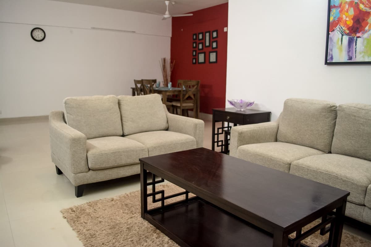 Una sala de estar con dos sofás beige, una mesa de centro de madera oscura, un tapete beige, una mesa de comedor con sillas y una pared de acento roja con cuadros enmarcados.