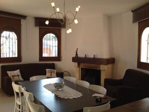 Comedor con mesa de madera, sillas blancas, chimenea y candelabro.