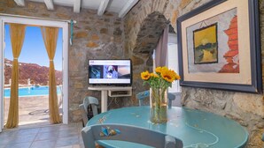 Een kamer met een stenen muur, een televisie, een tafel met een vaas zonnebloemen en uitzicht op een zwembad en bergen.
