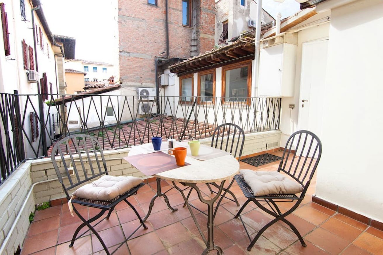 A small outdoor patio with a round table and four chairs, a white door, and a brick building in the background.