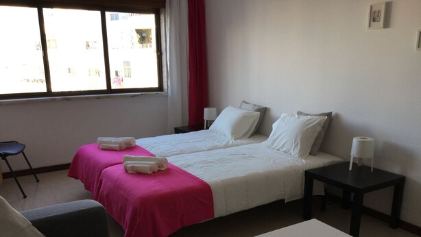 A hotel room with two beds, a small table, a chair, and a window with curtains. - APARTMENT LISBON - REBOLEIRA (metro / train) (Amadora)