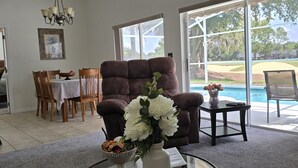 Un salon avec une table basse en verre, un fauteuil marron et un coin repas comprenant des chaises en bois et une nappe blanche.