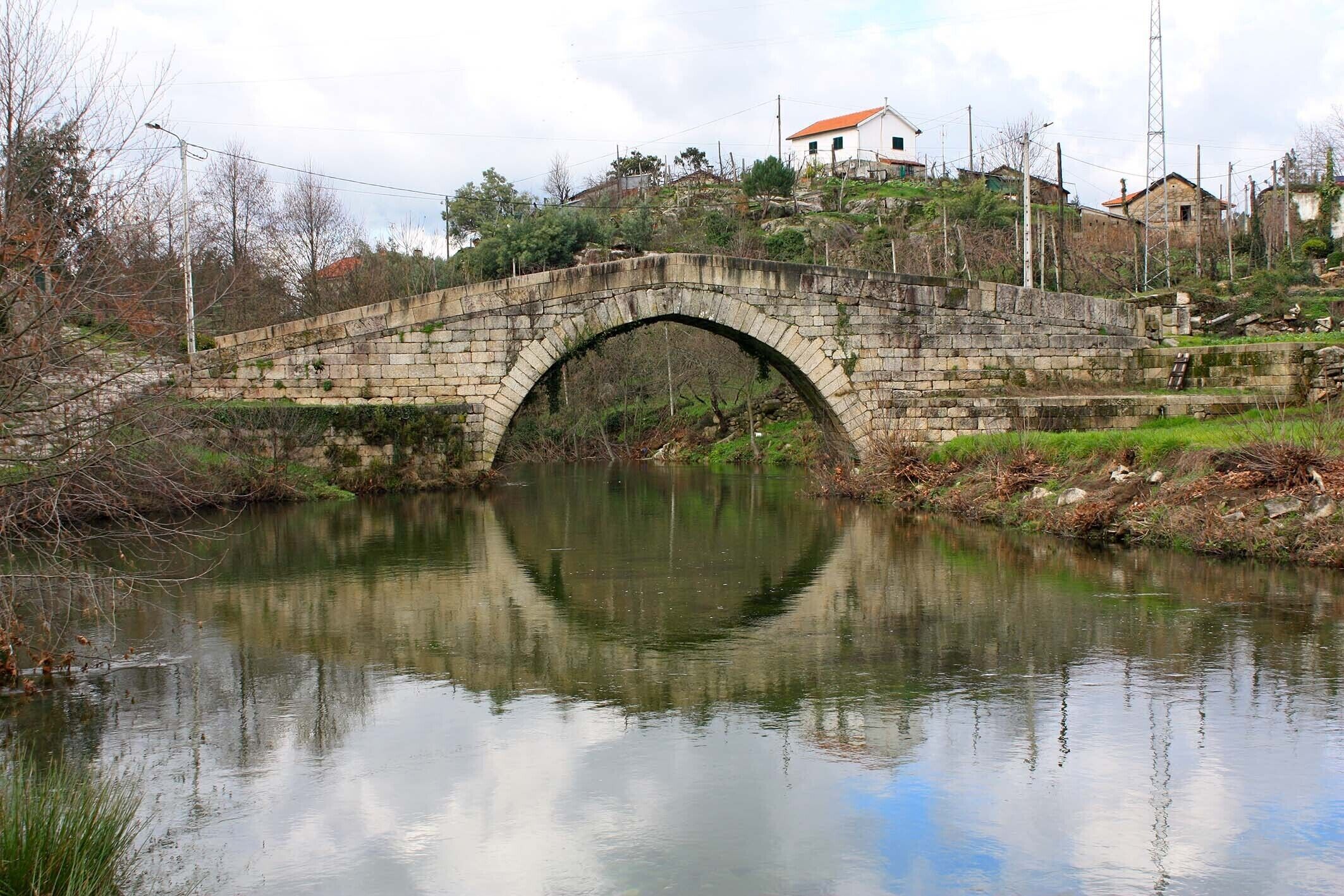 Un puente de piedra sobre un río tranquilo con reflejo, rodeado de vegetación y casas al fondo.