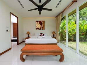 A hotel room with a bed, two bedside tables, a ceiling fan, a large window with a view of greenery, and a decorative wall piece.