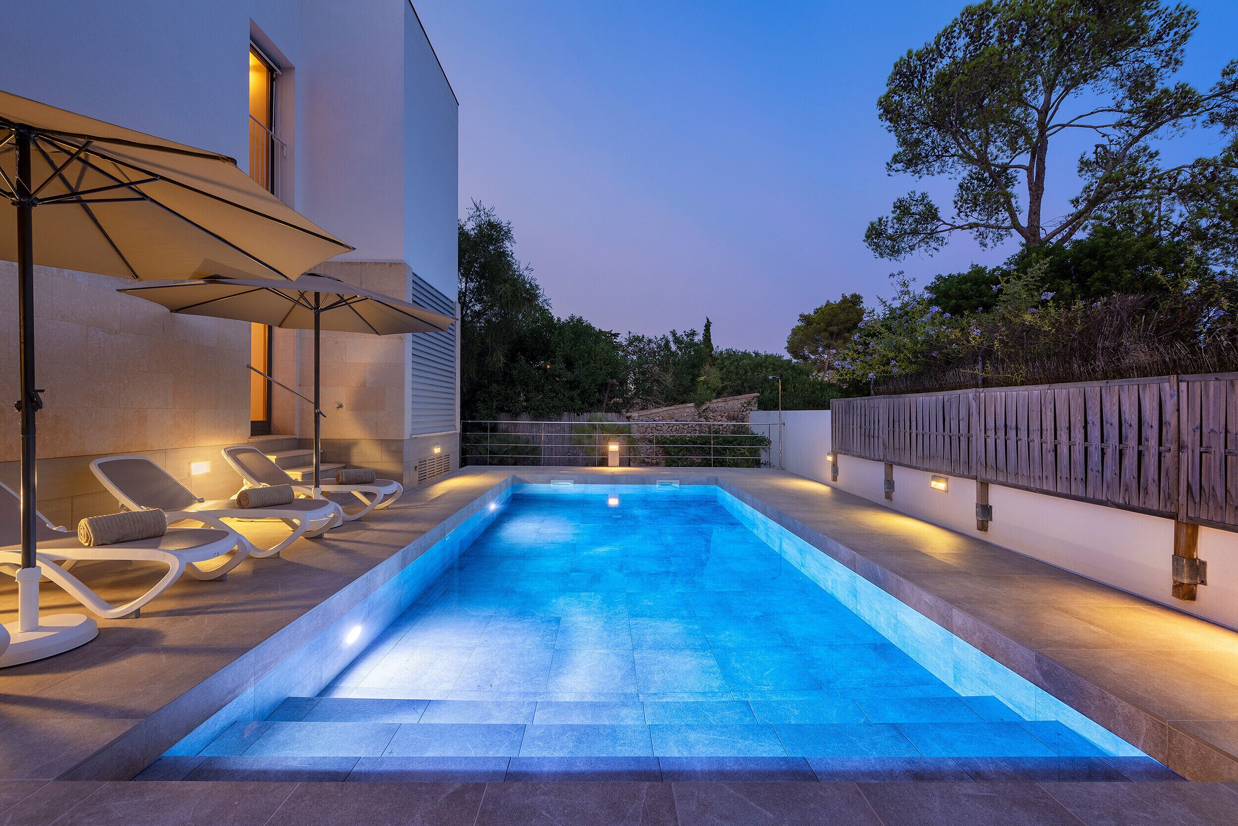 A swimming pool with lounge chairs and umbrellas, surrounded by a wooden fence and trees.