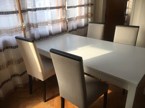 Un coin repas avec une table blanche et quatre chaises, un plancher en bois et une fenêtre ornée de rideaux légers.
