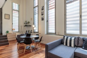 Una sala de estar moderna con un sofá gris, una mesa de comedor con sillas y ventanas grandes con persianas.