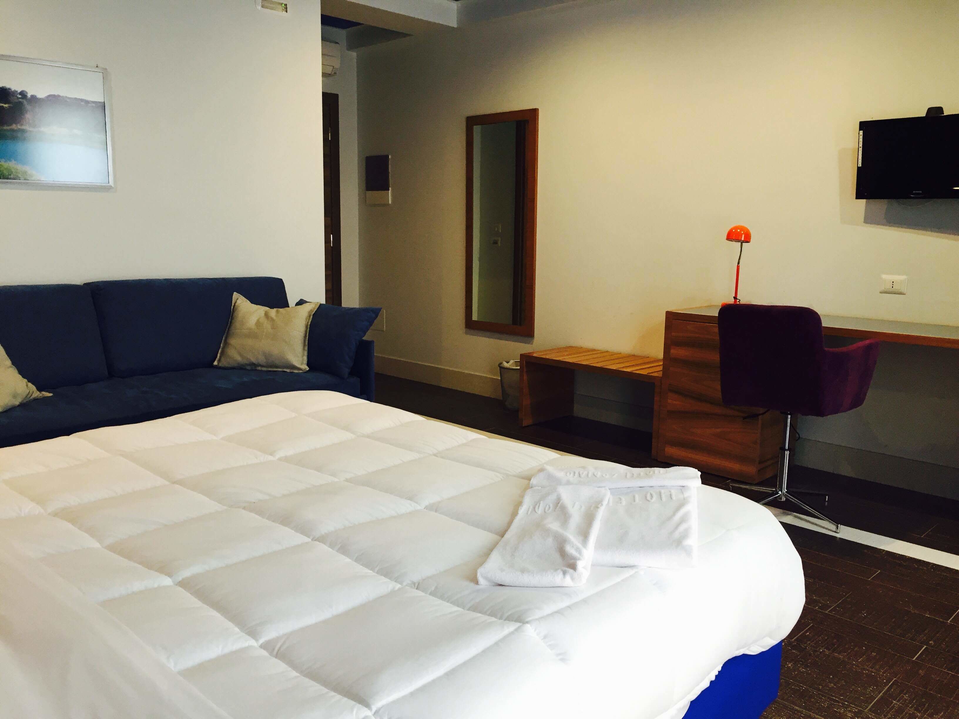 Ein Hotelzimmer mit einem großen Bett, einem blauen Sofa, einem Holztisch, einem Sessel, einem Fernseher und einem Spiegel.