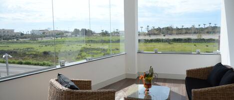 A balcony with wicker furniture, a glass table, and a view of a green landscape and buildings.