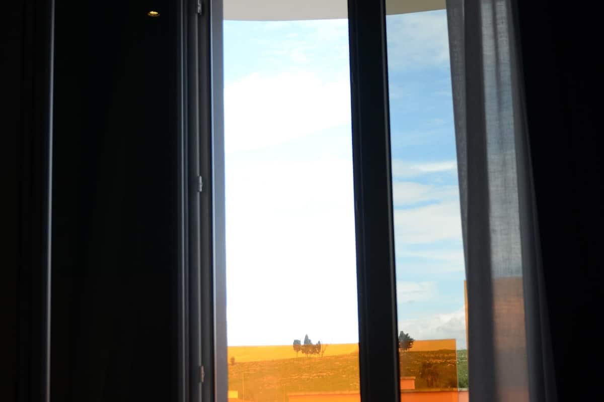 A room with a bed and a window. The window has a view of a field and a building in the distance.