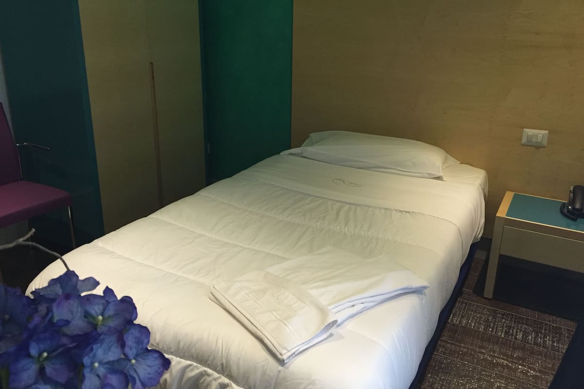 A neatly made bed with white bedding, a small bedside table with a lamp, and a chair against a green wall.