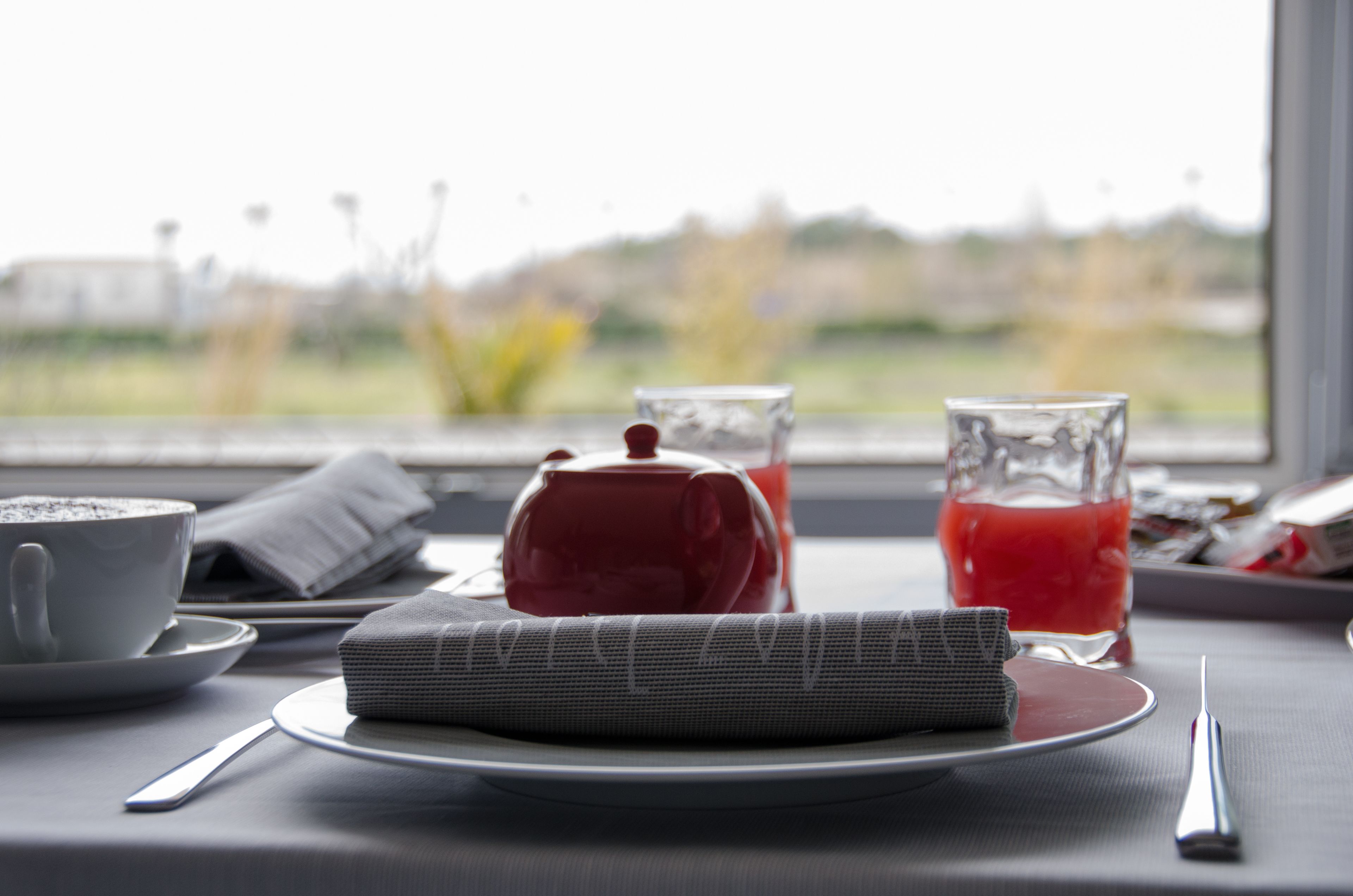 A breakfast setting with a teapot, two glasses of red beverage, a cup of coffee, and a folded napkin on a table by a window.