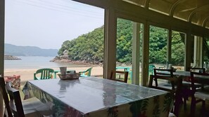 A restaurant with a view of a beach and forest. Tables are covered with plastic tablecloths, and there are chairs around them.
