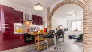 A modern kitchen with red cabinets, a wooden island, and a brick archway leading to a living area with a sofa and a TV.