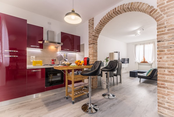 Una cocina moderna con armarios rojos, una isla de madera y un arco de ladrillo que da paso a una sala de estar con sofá y televisor.