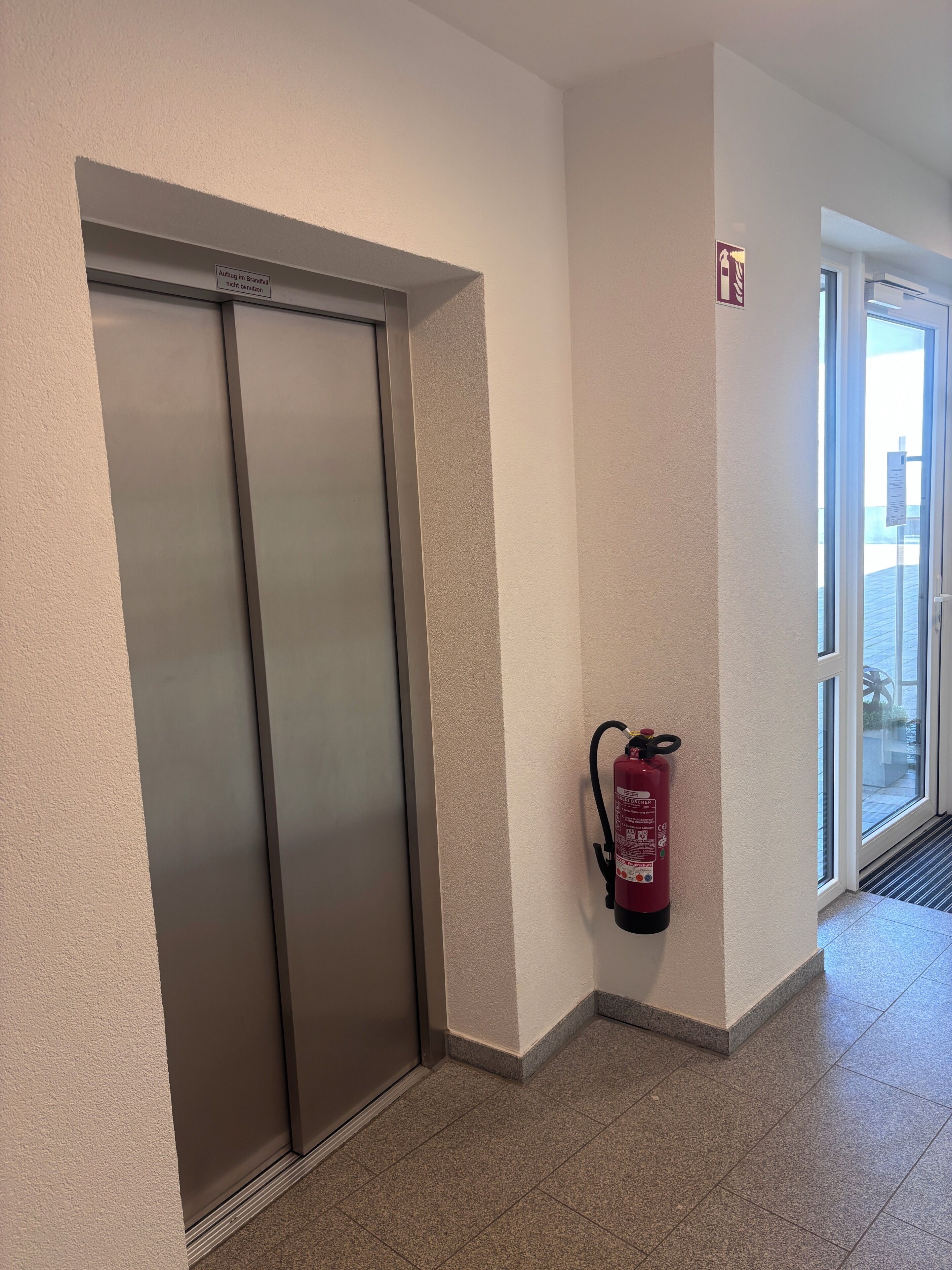 Un pasillo con un extintor colgado en la pared, una puerta y un ventanal.