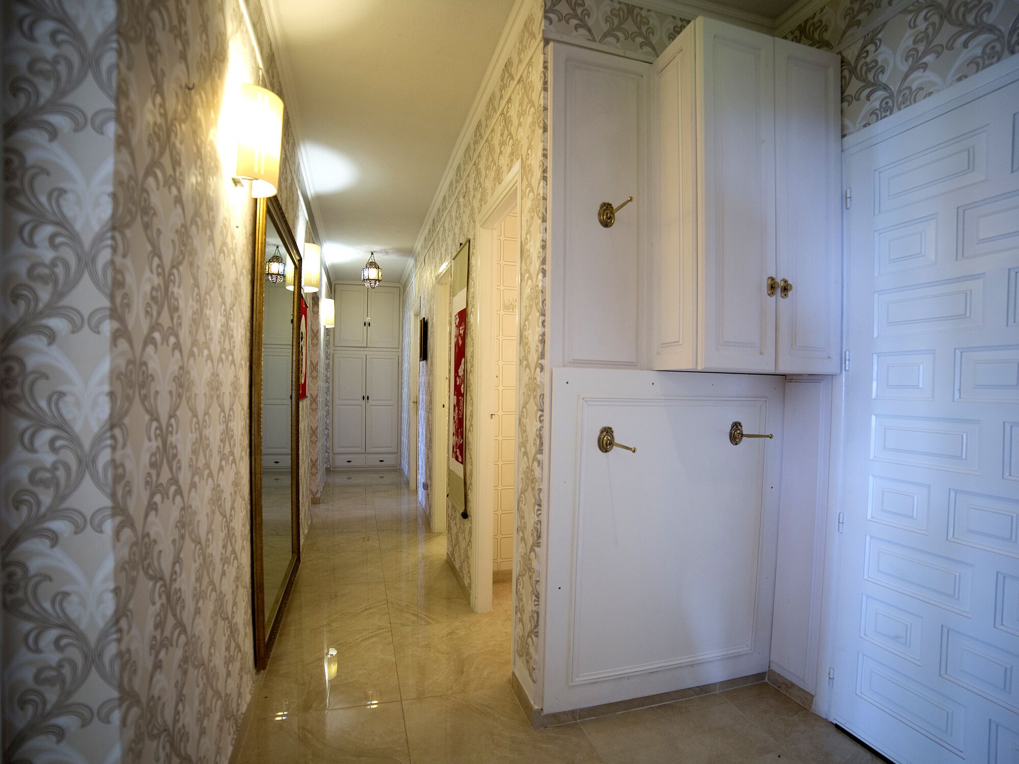 Un pasillo con papel tapiz estampado, un espejo y una puerta con ojo de cerradura.