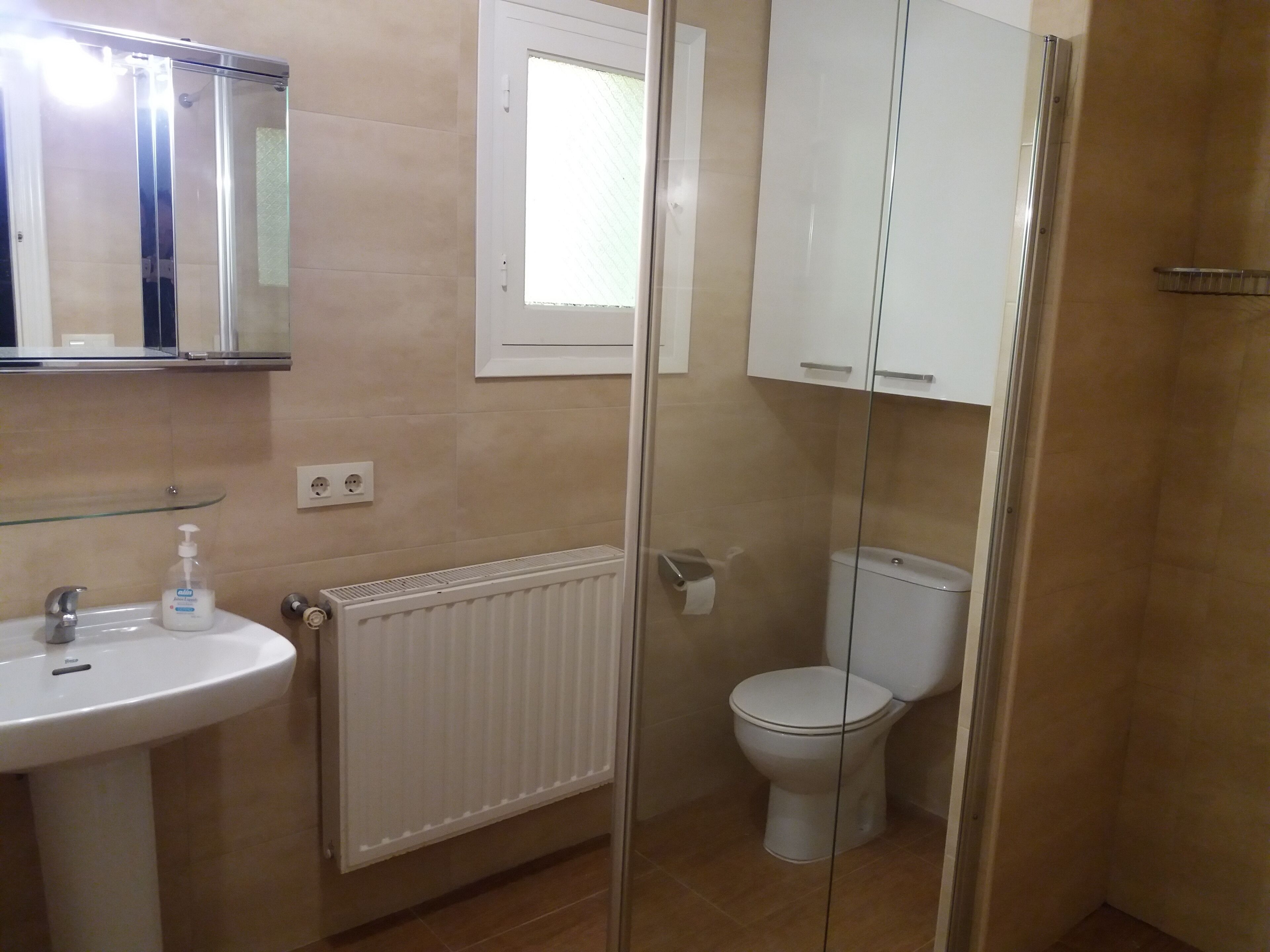 Un baño con cabina de ducha de vidrio, un lavabo blanco con dispensador de jabón, un inodoro y una ventana sobre el inodoro.
